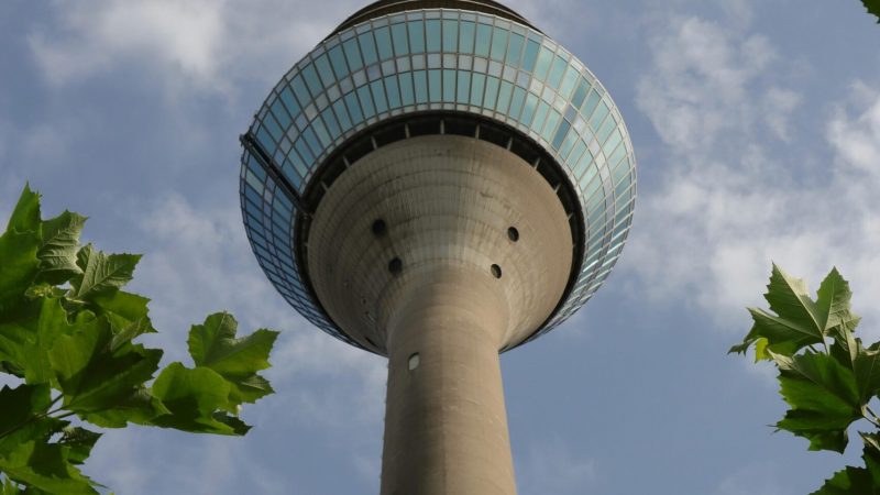 A tall concrete telecommunications tower with a round observation deck, seen from below through green leaves and blue sky, highlights connected jobsite.