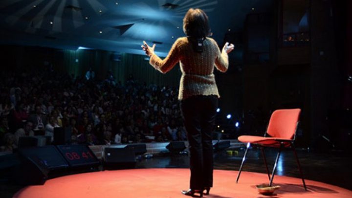 A person gestures on a red stage under bright lights, with a chair and basket nearby—capturing a connected jobsite presentation.