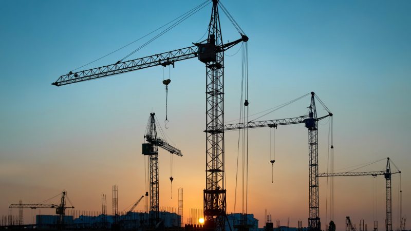 Construction cranes and unfinished buildings are silhouetted at sunset, highlighting a connected jobsite against a vibrant sky.
