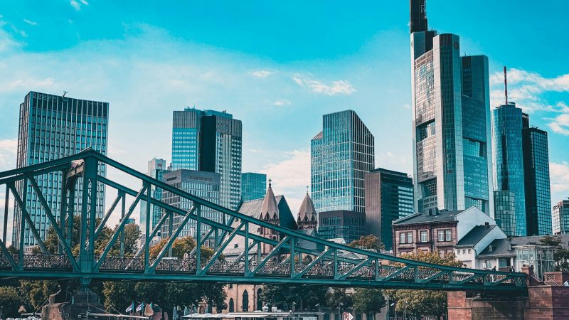 Modern city skyline with glass skyscrapers, Downtime Summit, iron bridge over river, historic riverside buildings; connected jobsite.