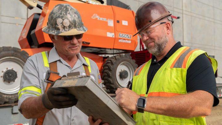 Two construction workers check digital documents on a clipboard at a site, with an orange lift and wall in the background, showcasing construction.