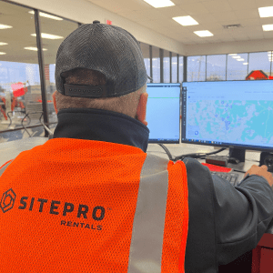 A person in an orange SITEPRO Rentals vest works at a multi-monitor desk showing maps, supporting construction equipment tracking.