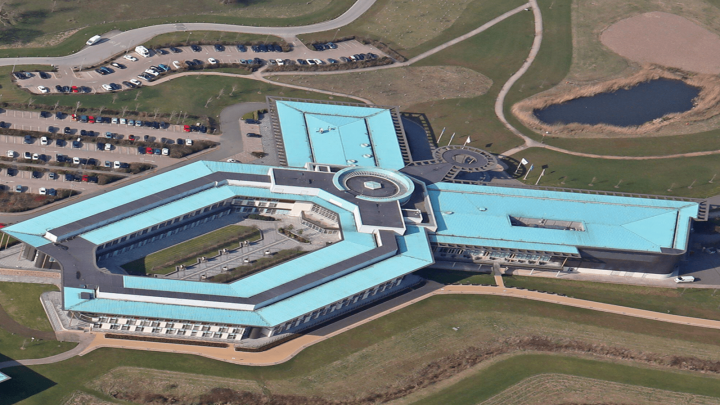 Aerial view of a modern building with a blue roof and geometric design, surrounded by green lawns, parking lots, and a small pond; ideal for connected jobsite.