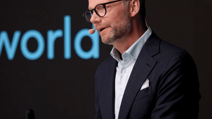 A man in glasses and a suit gestures while speaking on stage at the Trackunit Next event, related to construction IoT.