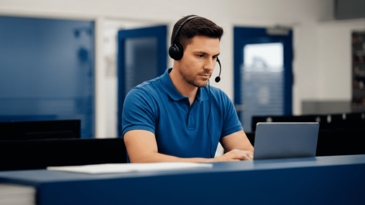 A man in a headset and blue polo works on a laptop at a modern office desk, reviewing fleet management data analytics.