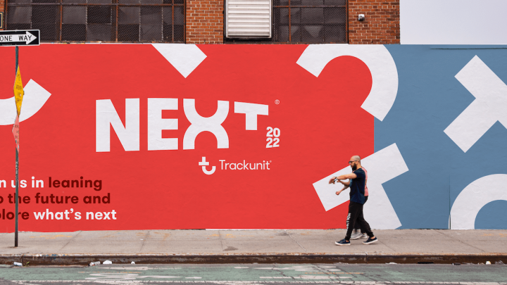 A person walks by a colorful city mural with bold hues and text about Trackunit NEXT 2022, referencing construction equipment tracking.