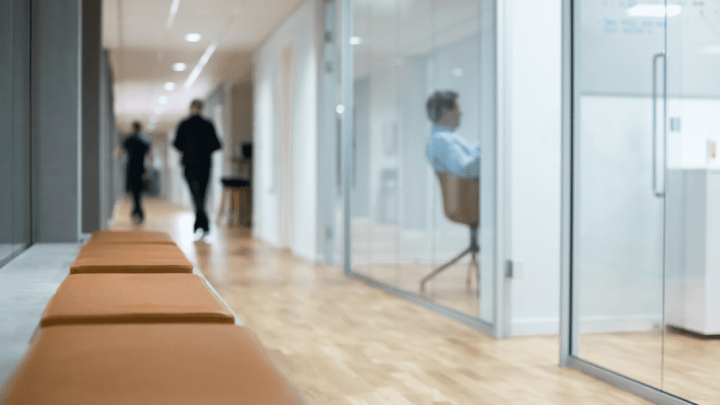 A modern office hallway with wooden floors, orange benches, glass-walled offices, and staff—suggesting a connected jobsite workspace.