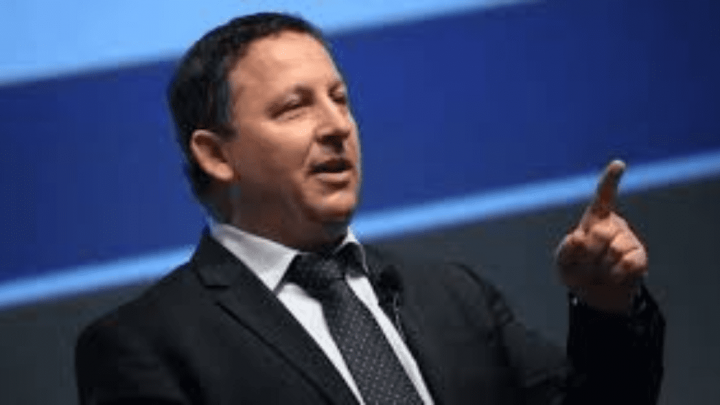 A chairman in a dark suit and tie gestures while speaking, pointing forward. Blurred blue background suggests a connected jobsite.