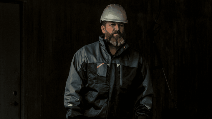 A bearded man in a white hard hat and dark jacket stands in a dim industrial space with telematics equipment in the background.