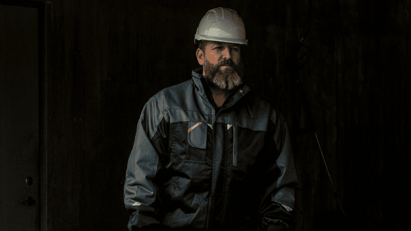 A bearded man in a white hard hat and dark jacket stands in a dim industrial space with telematics equipment in the background.