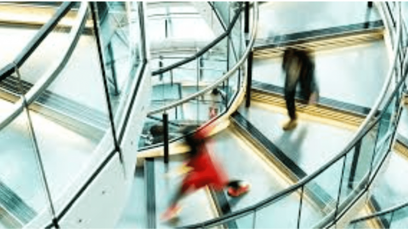 Blurred figures move on a spiral staircase with metal railings and glass panels, suggesting connected jobsite activity.