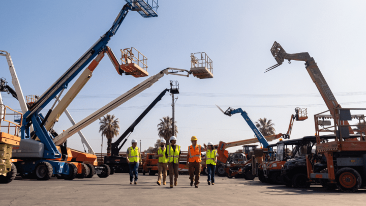 Five construction workers in safety gear walk among boom lifts on a lot, highlighting teamwork and a connected jobsite.