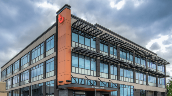 A modern three-story office building with glass windows, orange accents, and a red logo on the corner under a cloudy sky.