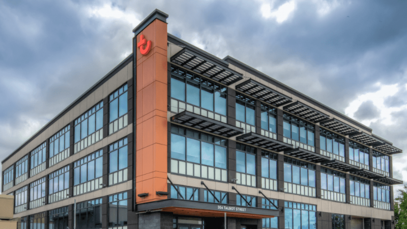 A modern three-story office building with glass windows, orange accents, and a red logo on the corner under a cloudy sky.