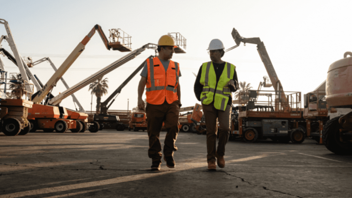 Two construction workers walk through a lot with boom lifts and equipment at CONEXPO, under a bright sky, representing construction.