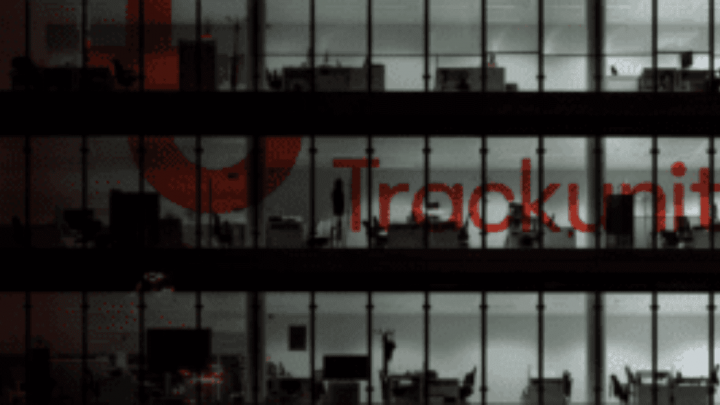 A dimly lit office building at night with workstations visible through large glass windows and the Trackunit logo on the facade.