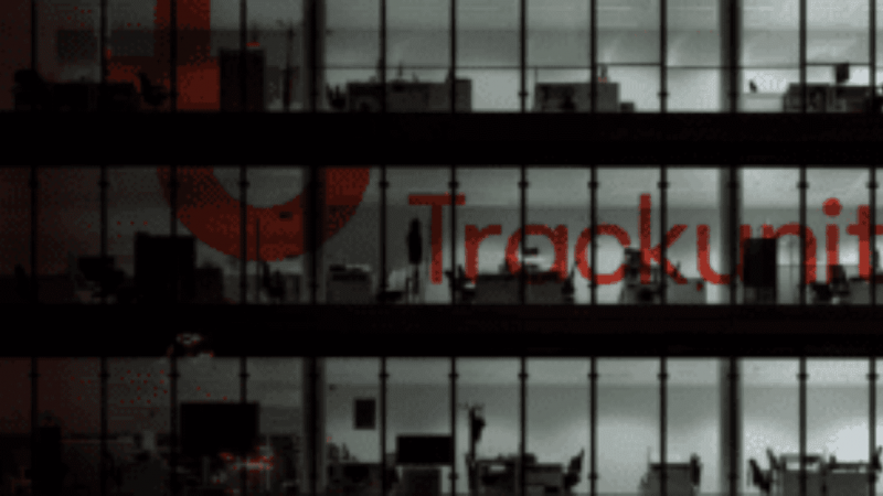 A dimly lit office building at night with workstations visible through large glass windows and the Trackunit logo on the facade.