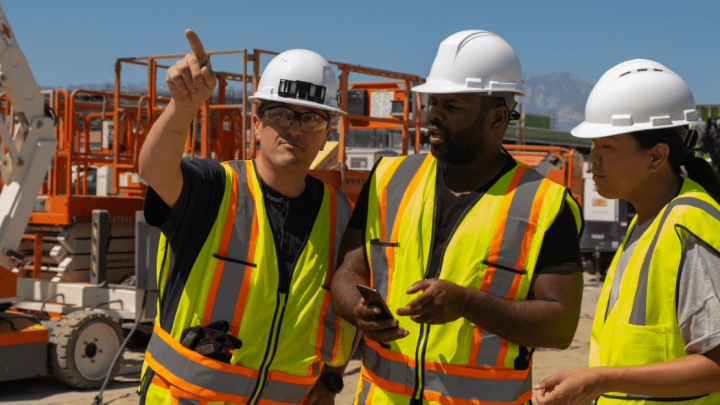 Three construction workers in hard hats and vests stand at a Conexpo site, discussing plans amid visible construction equipment.