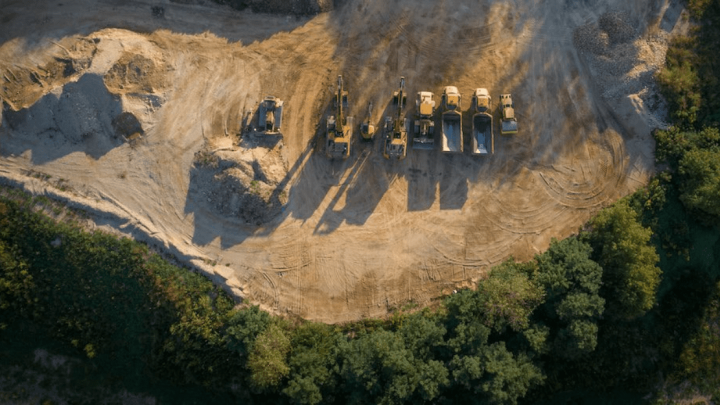 Aerial view of a construction site with excavators and trucks parked in a row, showing fleet management on sandy ground near trees.