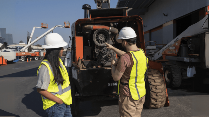 Two construction workers check heavy machinery with Trackunit on a connected jobsite, city skyline and equipment in the background.