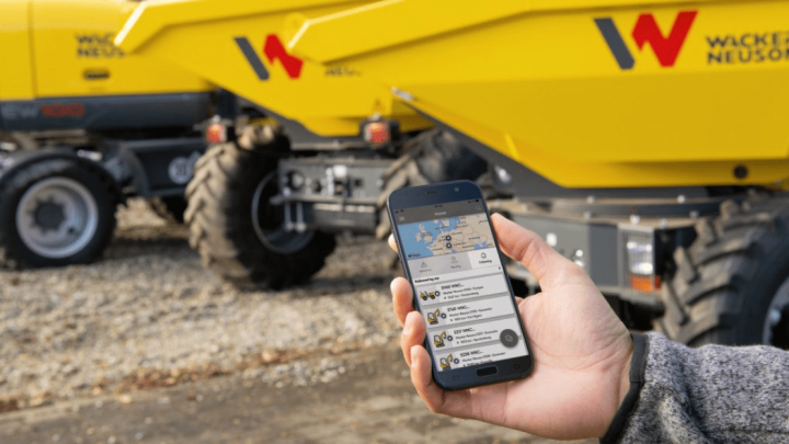 A person holds a smartphone with a Trackunit app in front of yellow Wacker Neuson equipment on a gravel lot, showing fleet management.