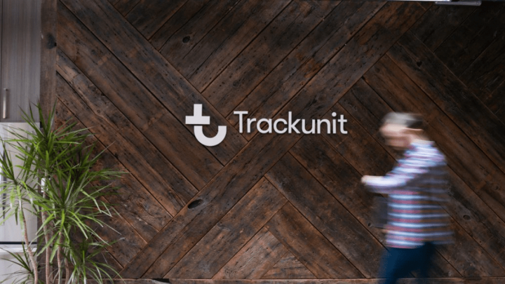 A blurred person walks by a dark wooden wall with the Trackunit logo; a green potted plant adds to the modern workspace.