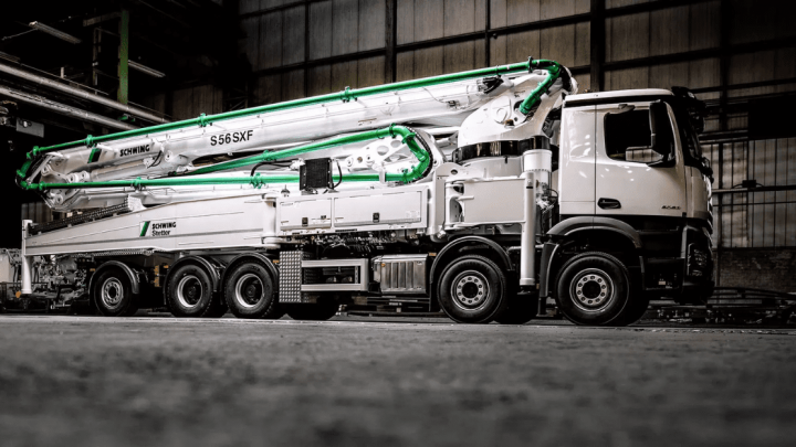 A white concrete pump truck with Trackunit and an extended boom is parked in a high-ceilinged warehouse for fleet management.