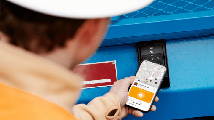 A person in a hard hat uses a smartphone showing a map and Trackunit location info beside blue construction equipment tracking panel.