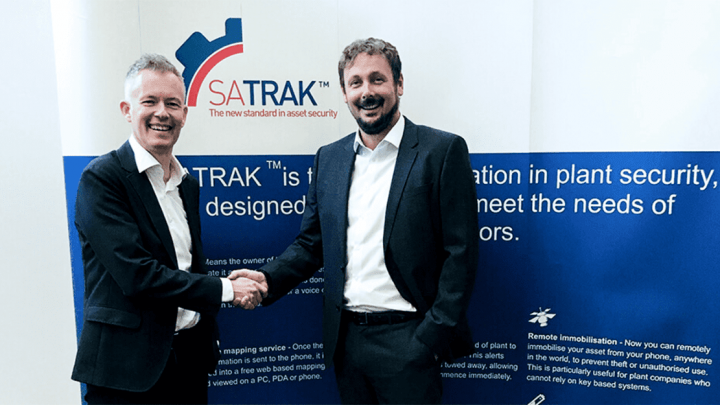 Two men in suits shake hands and smile in front of a blue and white SATRAK™ banner for telematics and construction equipment tracking.