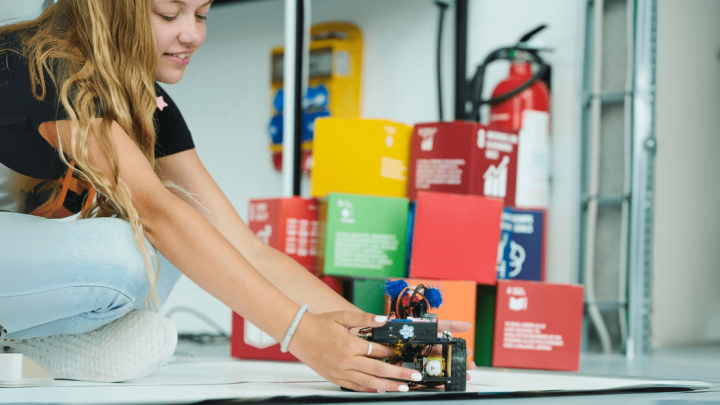 A girl kneels, smiling as she adjusts a small robot. Colorful Trackunit boxes hint at construction IoT in the background.