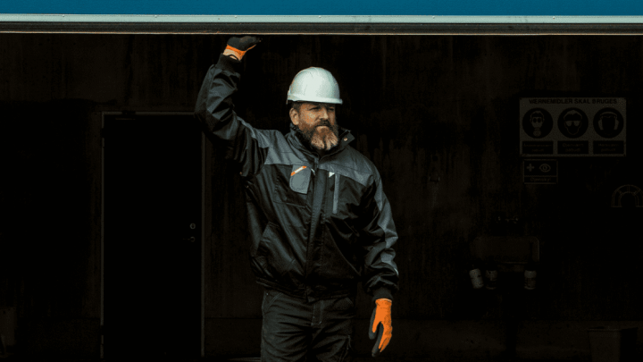 A man in a white hard hat and orange gloves stands outside, reaching for a ledge. Construction IoT safety signs are on the wall behind him.