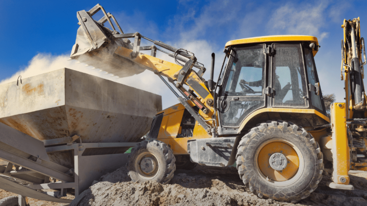 A yellow backhoe lifts a concrete block at a sunny, dusty construction site, highlighting modern construction and equipment tracking.