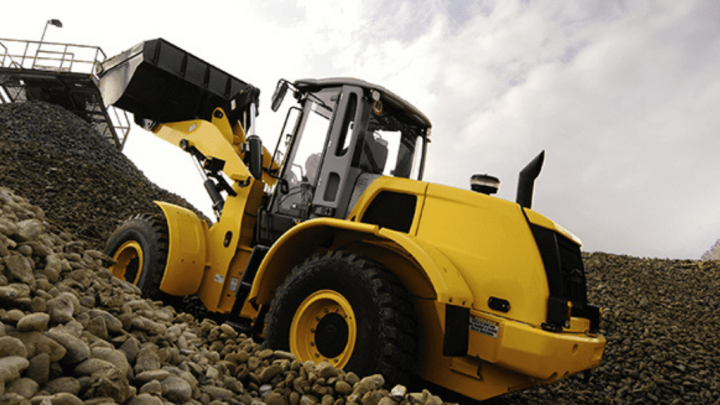 A yellow front-end loader with telematics lifts rocks at a construction site under a cloudy sky.