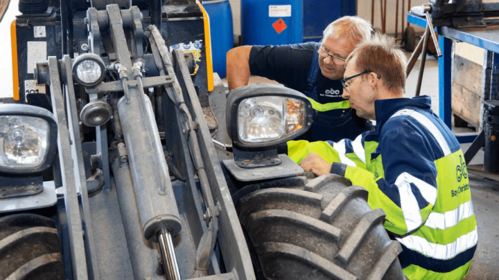 Two men in work uniforms inspect a large industrial vehicle in a digital workshop, highlighting construction equipment tracking.