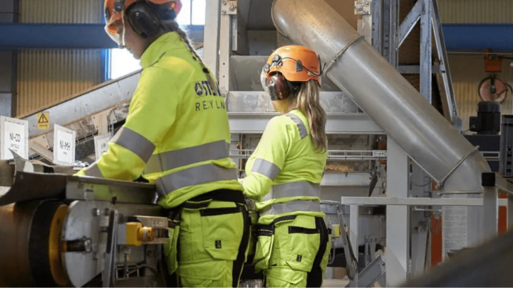 Two workers in high-visibility gear operate digital machinery at an industrial site, with conveyors and pipes, showing a connected jobsite.