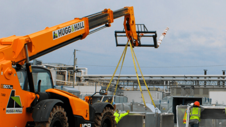A telescopic forklift lifts a metal object as three workers guide it at a construction site with digital equipment, showing a connected jobsite.