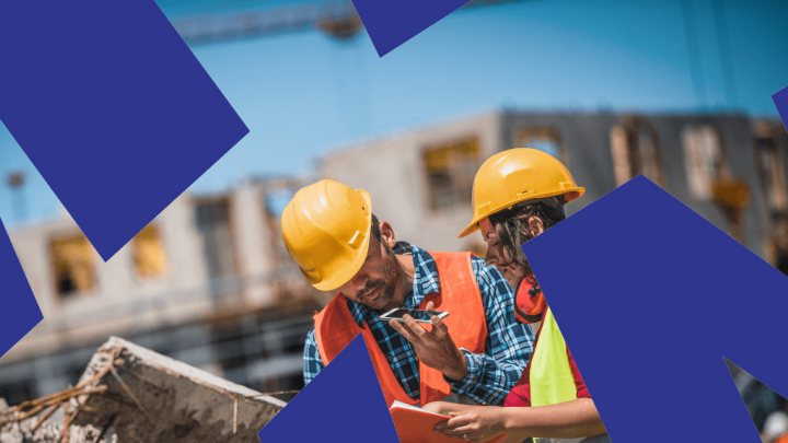Two construction workers in safety vests and helmets review documents at a building site, highlighting a connected jobsite.