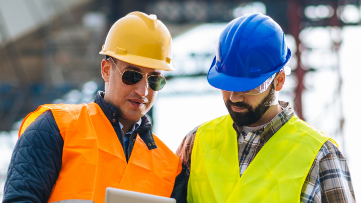 Two construction workers in safety vests and hard hats discuss telematics on a tablet at a construction site.
