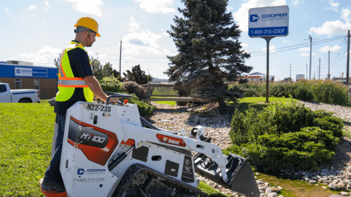 A construction worker uses a mini track loader with digital controls near a Cooper Equipment Rentals sign, showing connected jobsite tech.