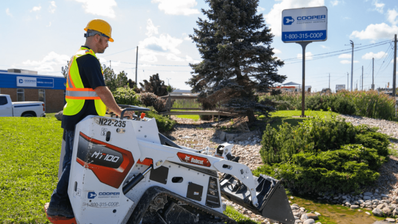 A construction worker uses a mini track loader with digital controls near a Cooper Equipment Rentals sign, showing connected jobsite tech.