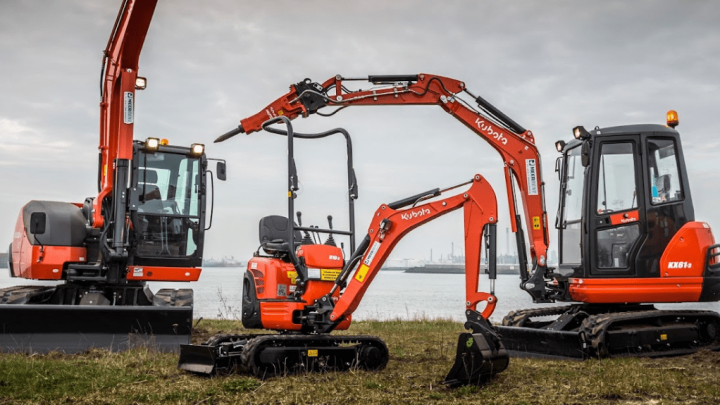 Three orange excavators of varying sizes parked on grass by water, with industrial buildings nearby, showing a connected jobsite.