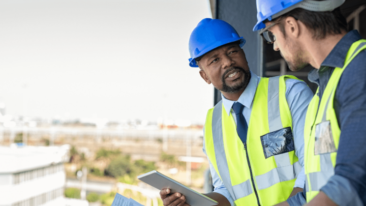 Two construction workers in safety vests and helmets discuss digital plans on a connected jobsite, with buildings in the background.