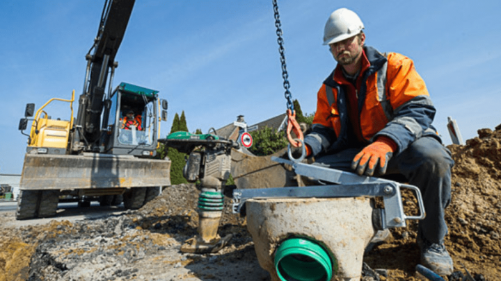 A construction worker lifts a drainage pipe with a chain at a site, referencing digital plans on a tablet for connected jobsite tasks.