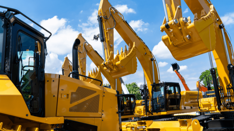 Large yellow excavators with telematics are lined up outdoors under a blue sky, their arms raised against trees in the background.