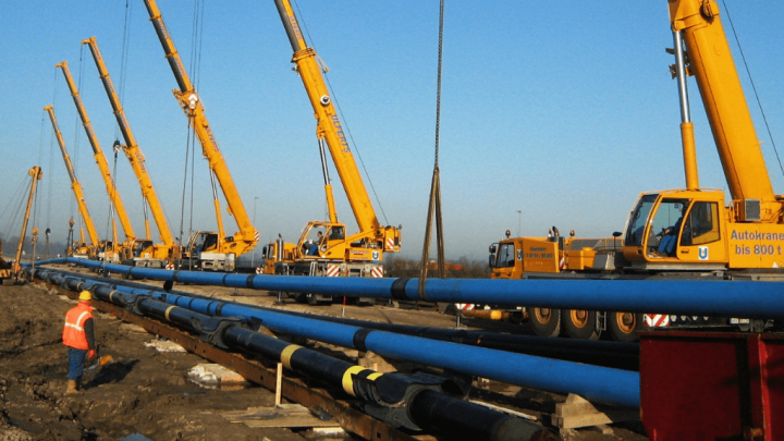 Yellow cranes lift blue pipes at a construction site as workers with digital tools oversee the connected jobsite on a clear day.