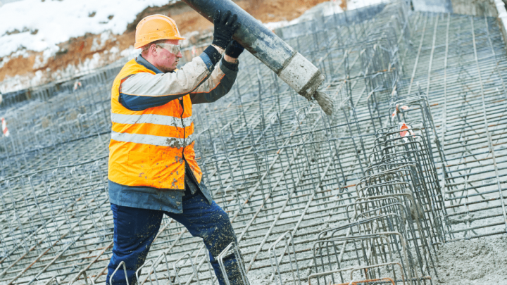 A construction worker pours concrete onto rebar at a connected jobsite on a snowy day, wearing safety gear.