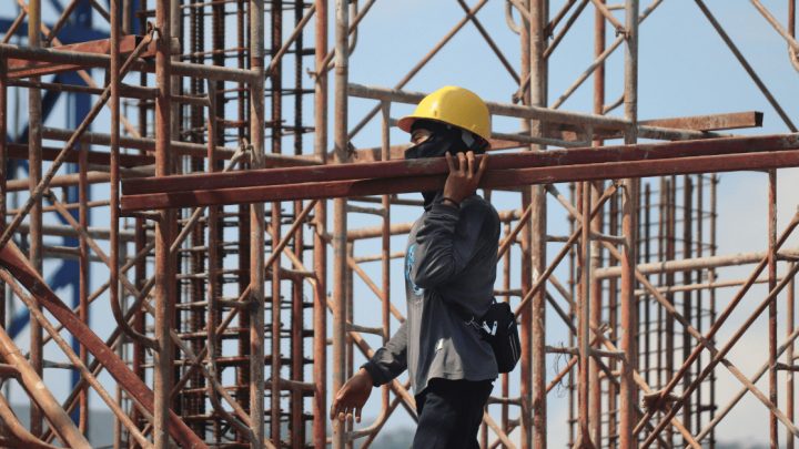 A construction worker in a yellow hard hat carries a metal beam on a connected jobsite with scaffolding and steel structures.