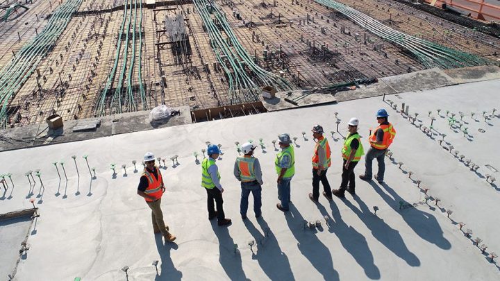 Six construction workers in safety vests and helmets discuss on a building site with rebar, cables, and concrete, showing a connected jobsite.