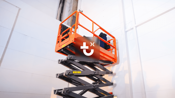 A bright orange scissor lift is raised in a large, white industrial space, showing construction equipment tracking in action.