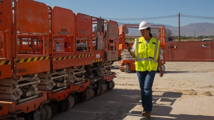 A worker in a hard hat and safety vest inspects orange scissor lifts outdoors, with mountains behind, illustrating construction equipment tracking.
